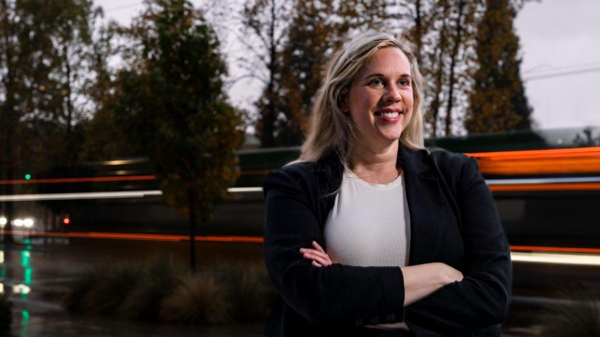 Portrait of a woman with long blond hair and a blue jacket standing outside as the blurred lights of passing traffic pass by.