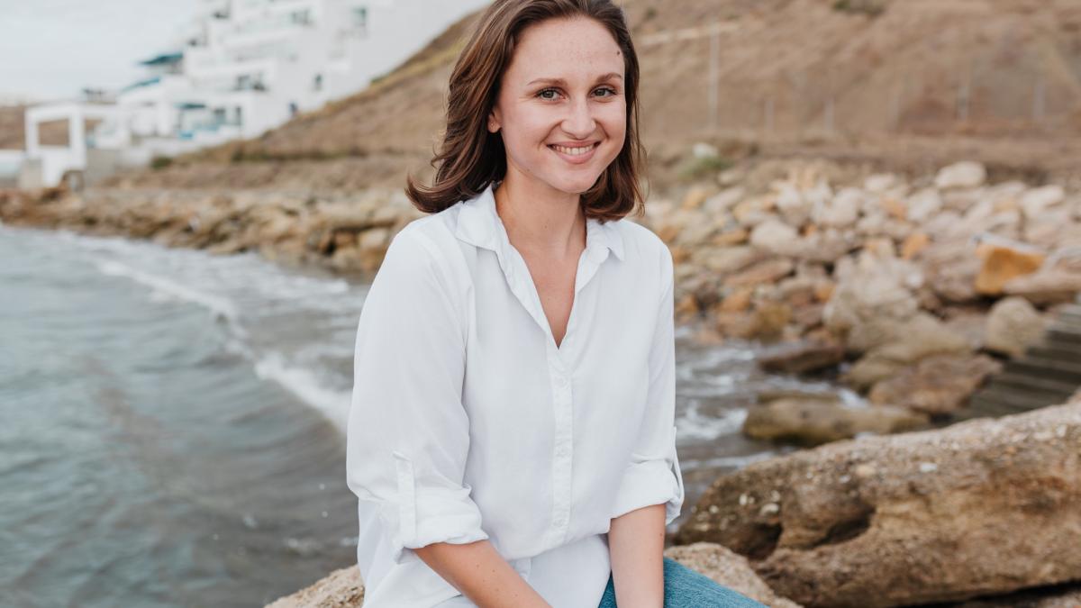 A young woman in a white shirt and jeans poses along a marine waterfront.