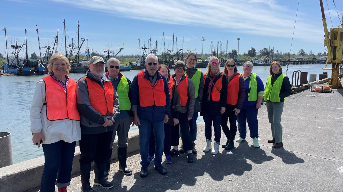 A group of people pose for a photo near a fishing harbor. They each wear orange safety vests.