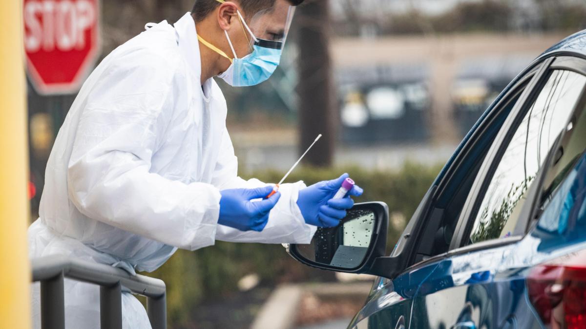 A man in a face mask and protective gear holds a tube and cotton swab near the driver's side window of a car.