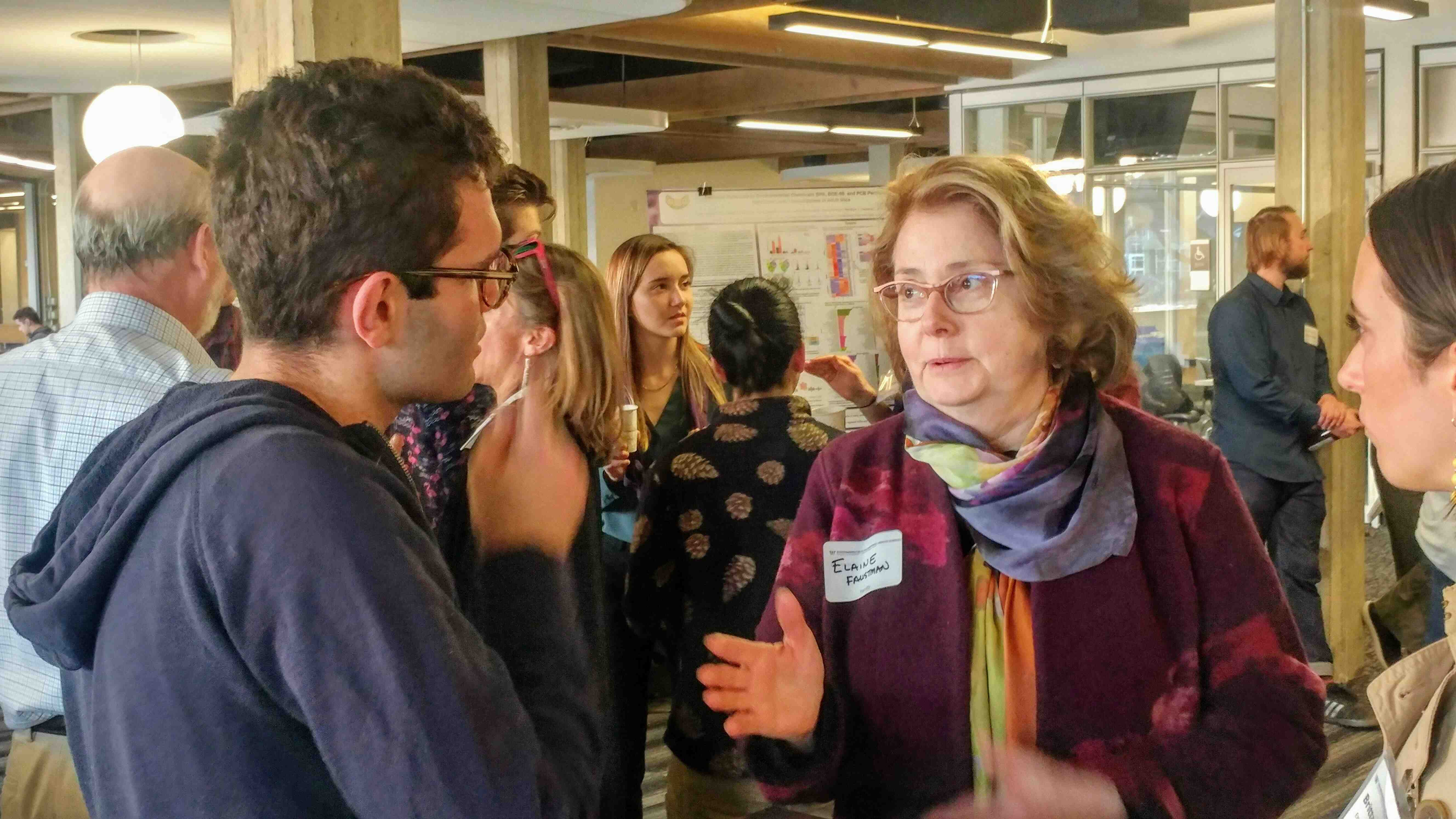 Elaine Faustman (on right) speaks with a student (on left) at a poster session with other people in the background.