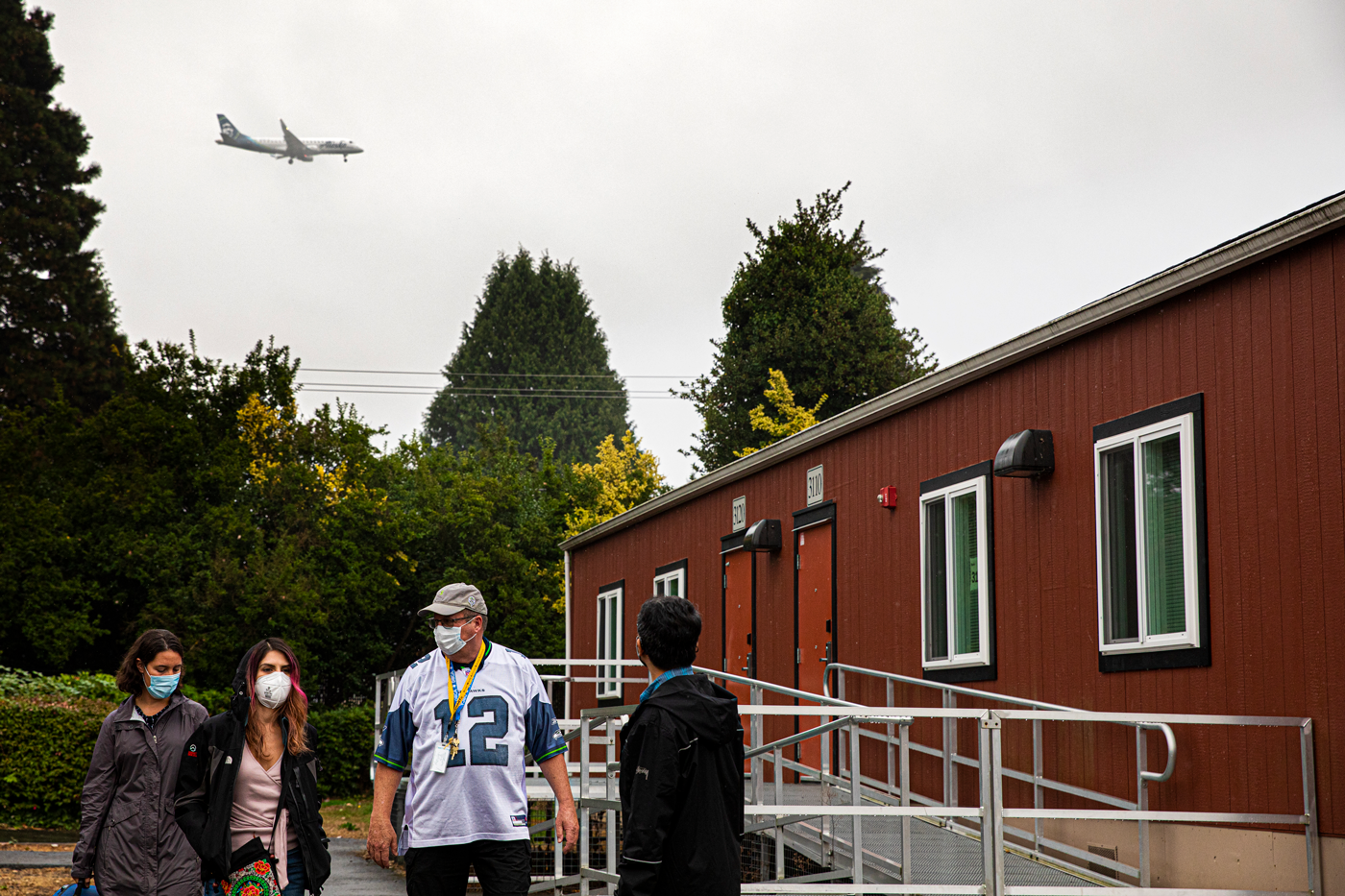 Four people stand outside a modular school building with a plane flying overhead.