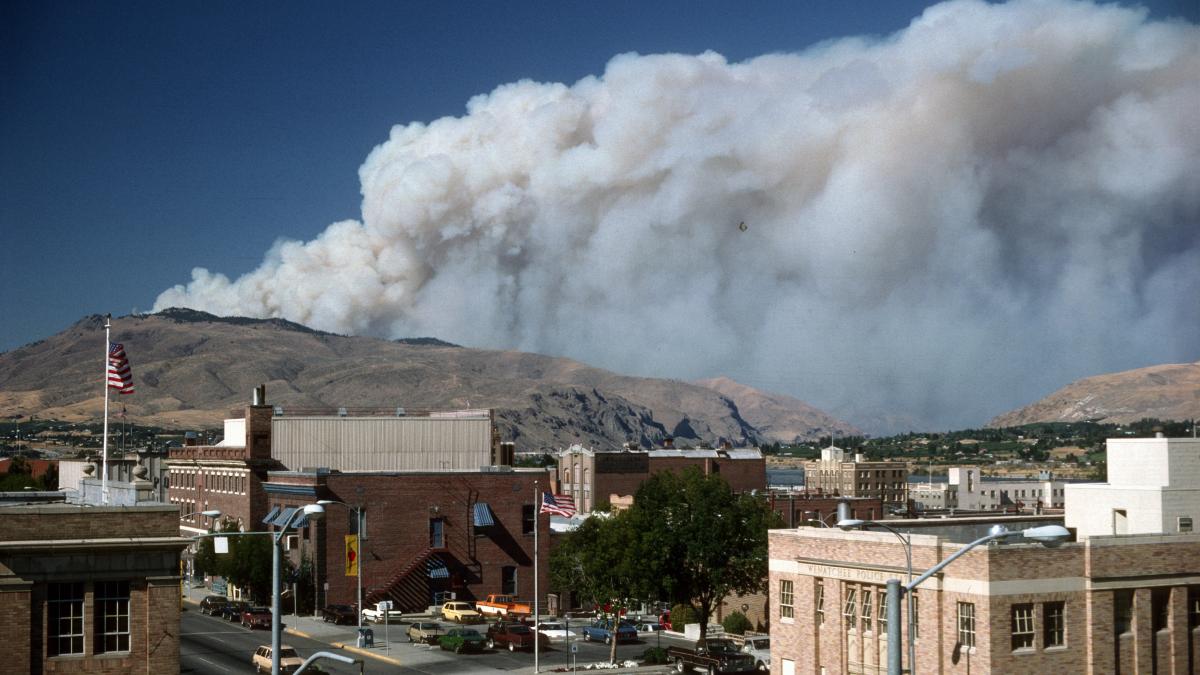 A plume of wildfire smoke behind the center of a small town showing brick buildings and cars. 