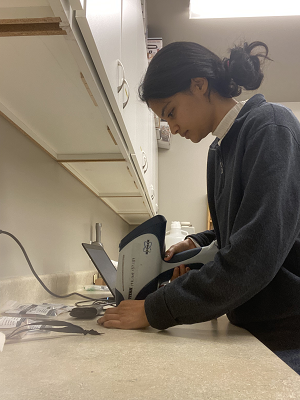 Mokashi stands at a lab bench holding an X-ray fluorescence analyzer over an eyeliner sample.