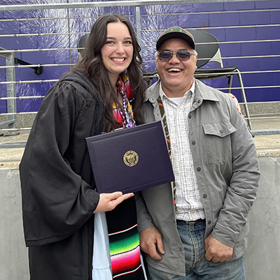 Belen Salguero stands in graduation gown holding a diploma next to her father.