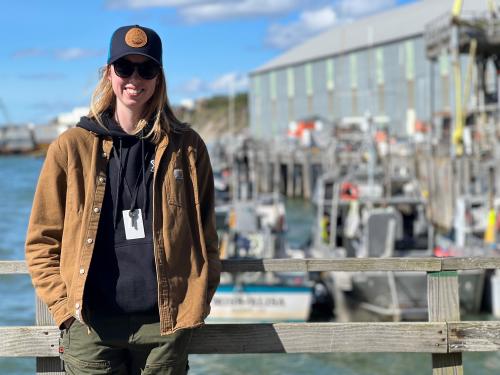 A woman in a ball cap and sunglasses smiles while standing near water.