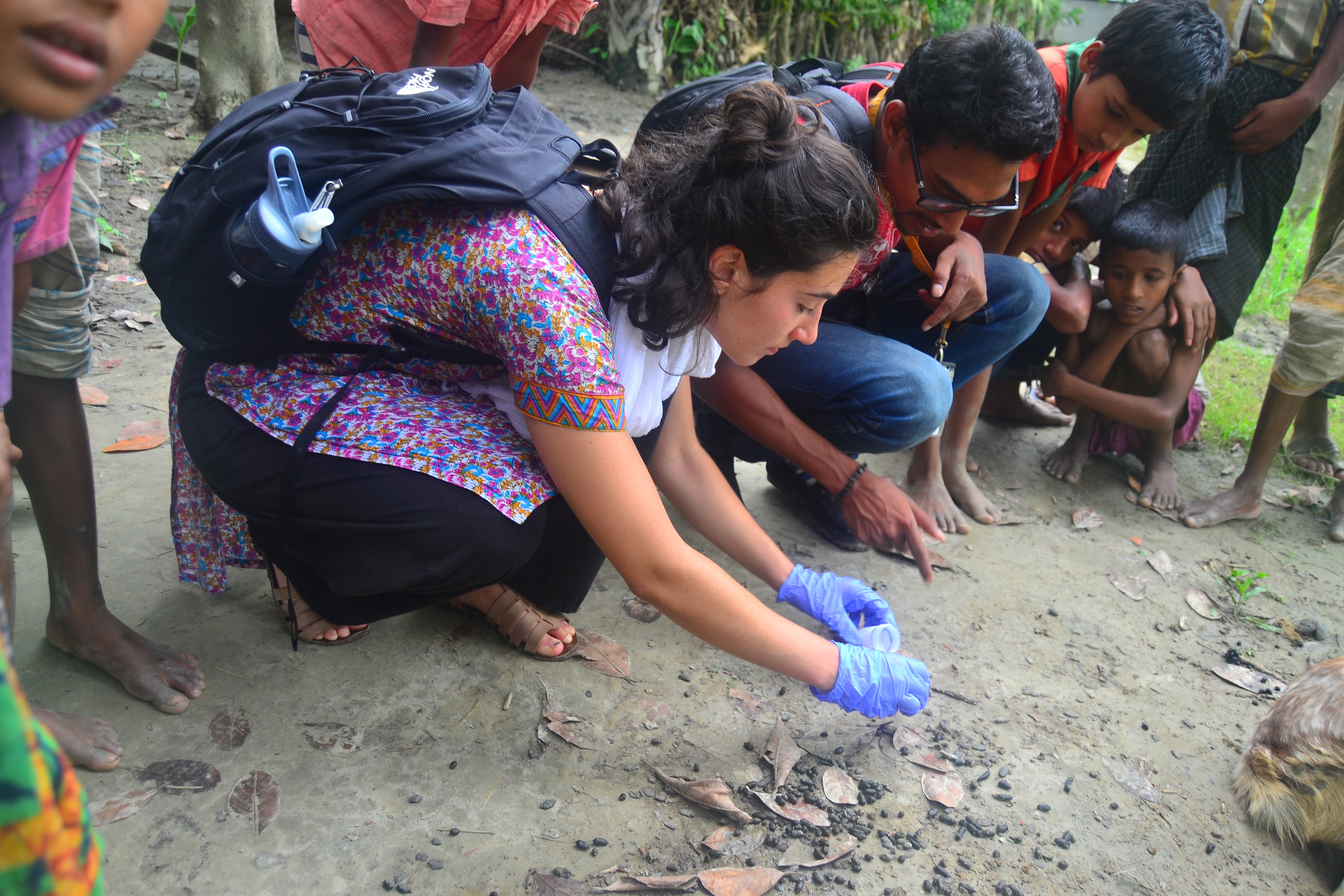 A woman wearing surgical gloves kneels on the ground next to a group of people while collecting goat poop.