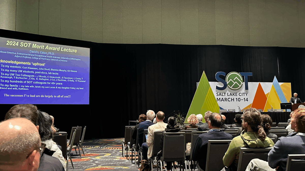 Eaton stands at a podium addressing an auditorium of people next to a sign reading SOT Salt Lake City March 10-14. A powerpoint slide with acknowledgments is up on the presentation screen.