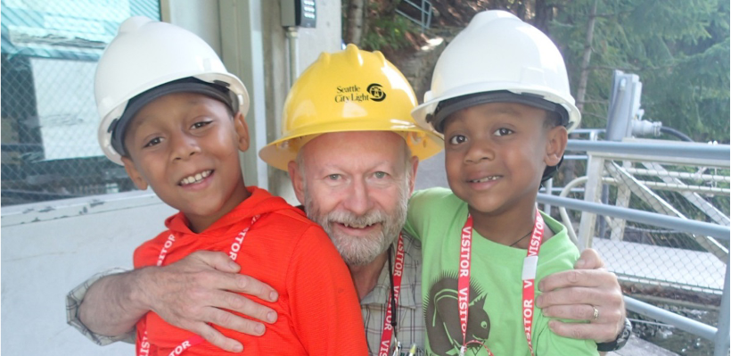 A man kneels down between two children, all of whom are wearing safety helmets.