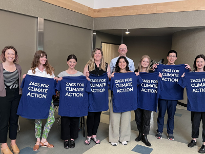 A group of 9 people stands together, with some of them holding blue T-shirts reading "Zags for Climate Action."