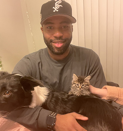 A man wearing a baseball cap holds a kitten and sits next to a dog.