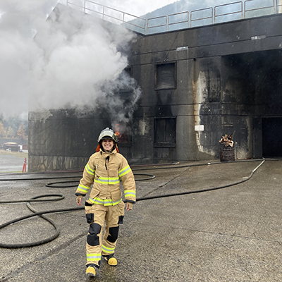 A woman stands in firefighter turnout gear in front of a burned building.