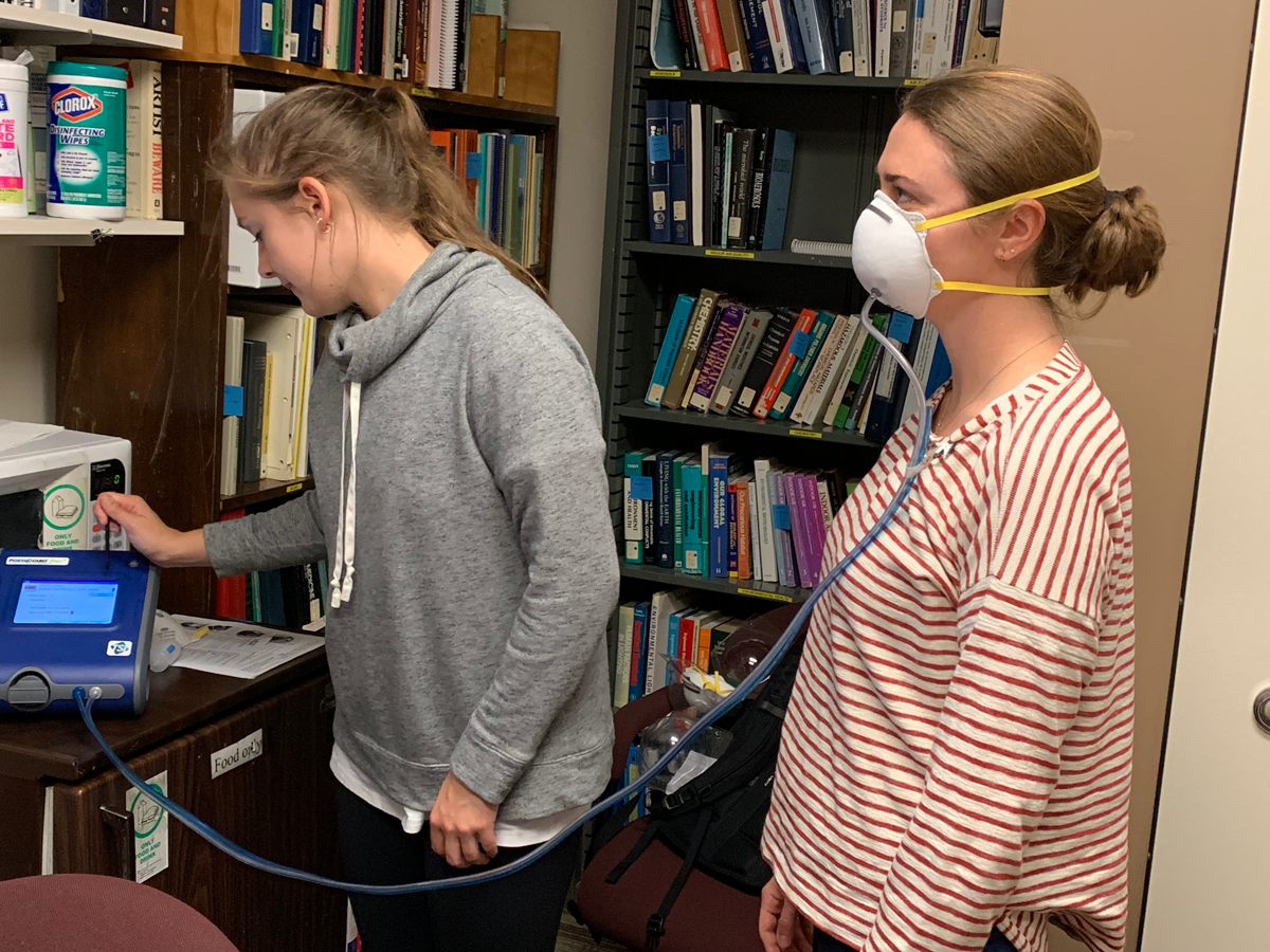 A woman checks the fit of a face mask respirator on another woman.