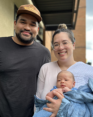 Shelley and her partner, a man in baseball cap, stand outside together as Shelley holds their infant son.