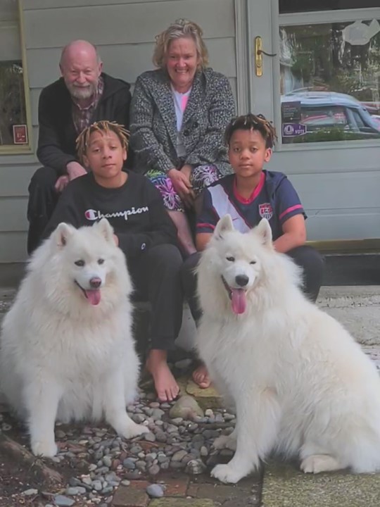 A man poses on his porch surrounded by his wife, grandsons and two dogs.