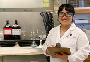 Woman in lab coat and glasses standing in front of a fume hood.