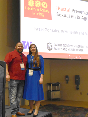 Drury stands next to a man in front of a projector screen showing a slide from the Basta project. 