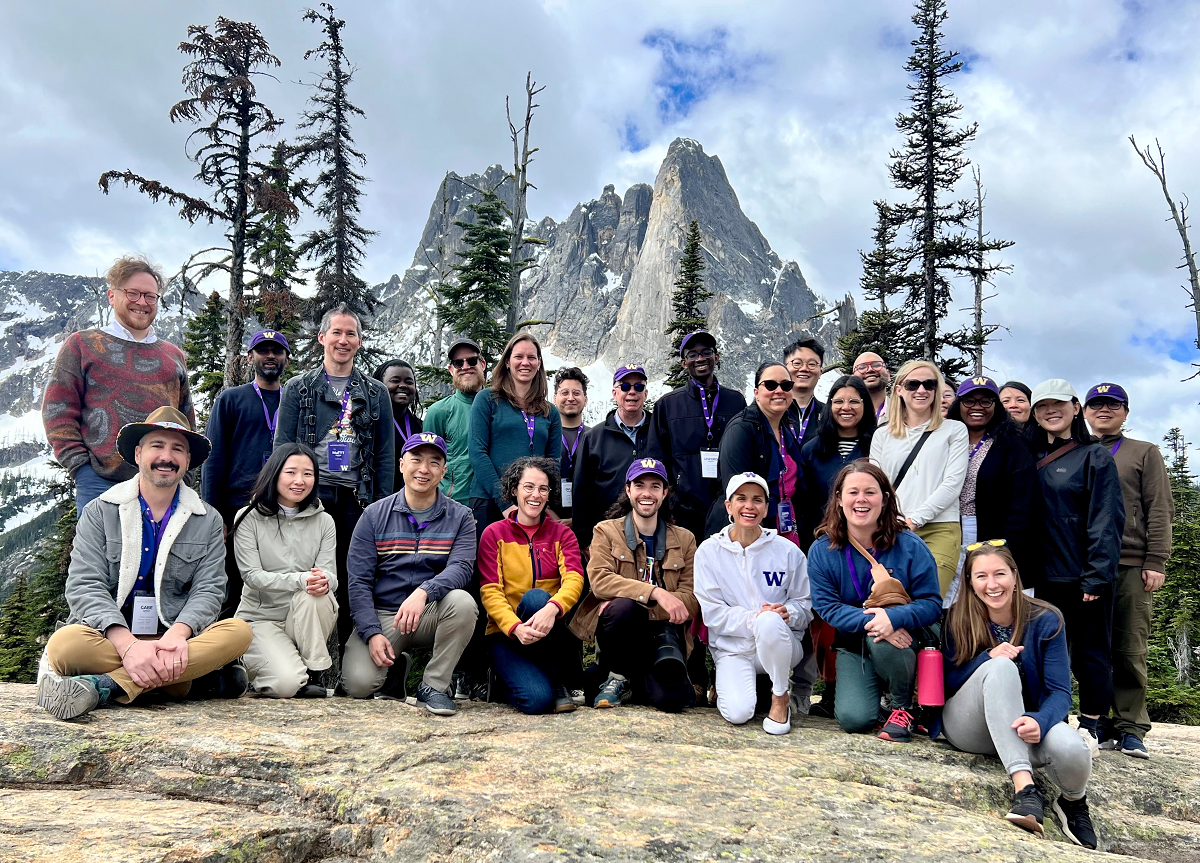 About 30 people stand in rows for a group photo in front of steep mountain peaks.