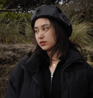 A young woman in a black beret and jacket poses against a hillside.