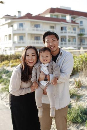 A man and woman pose outdoors along with their baby boy, with a large house framed in the background.