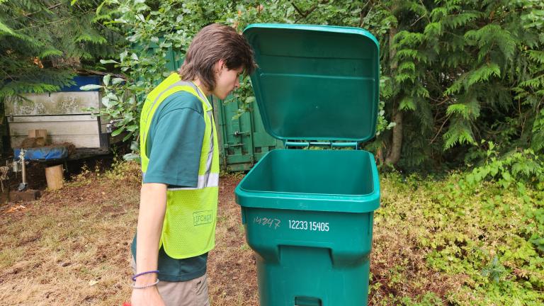 Kaidan St. Louis stands in a safety vest looking into a recycling bin in a backyard while holding it open.