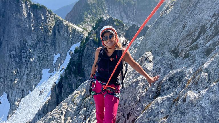 Woman in a climbing harness with rope stands on a mountain peak with mountains and snow in the background.