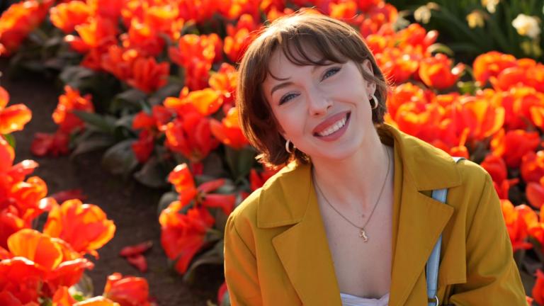 Ally Clonch smiles wearing a yellow coat in front of a field of orange tulips.