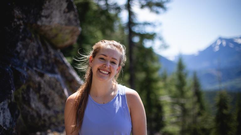 A woman in a purple shirt smiles against an outdoor backdrop of trees and mountains.