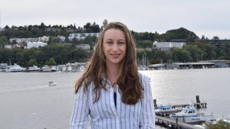 A woman with long hair and a striped shirt smiles while standing near a body of water.