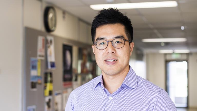 A man wearing glasses and a purple checked shirt poses in a hallway.