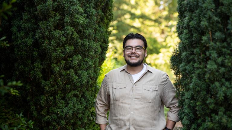 A man with glasses and a tan shirt poses between two evergreen shrubs.