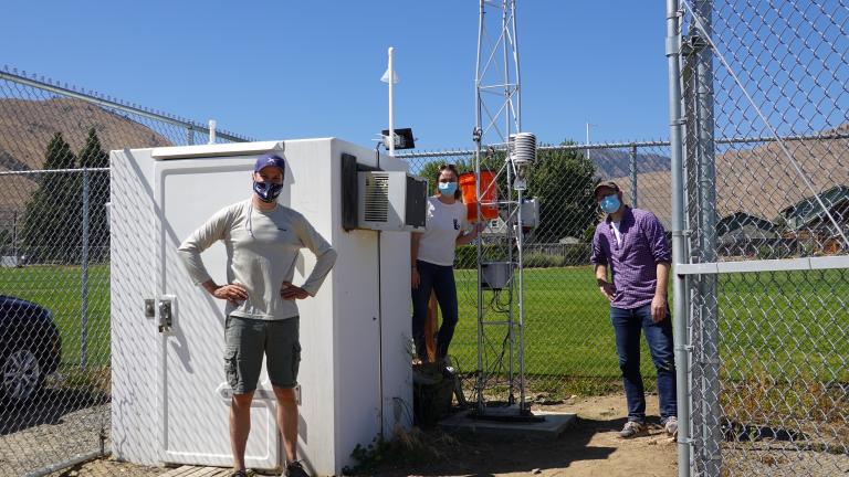 3 people wearing face masks stand next to weather monitoring equipment inside a chain-link fence.