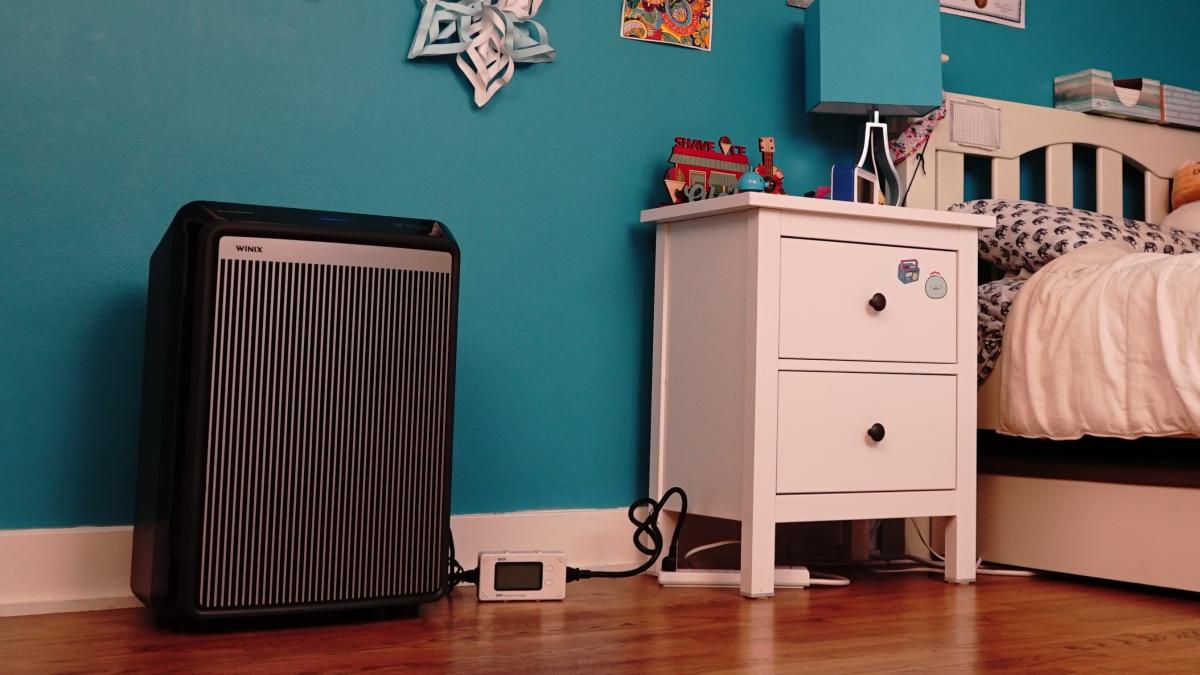 A child's bedroom scene with teal walls, paper decorations on the walls, a white bedframe on the right, a white nightstand in the center with a teal and silver lamp, and a black rectangular Winix air cleaner on the left.