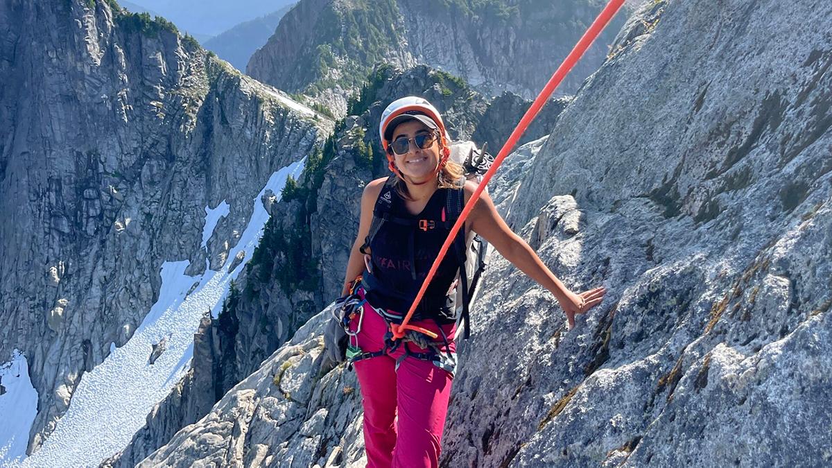 Woman in a climbing harness with rope stands on a mountain peak with mountains and snow in the background.