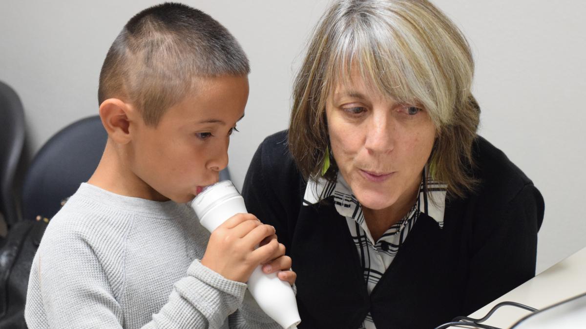 A young boy in a gray sweater breathes into a device measuring lung function, and a woman next to him in a black sweater demonstrates how to breathe while checking a computer monitor.