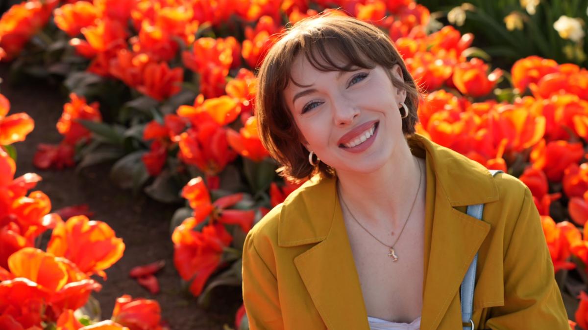 Ally Clonch smiles wearing a yellow coat in front of a field of orange tulips.