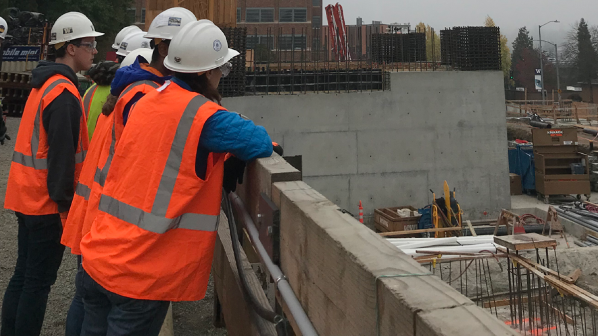 A group of UW students in orange vests stands by a wall looking into a construction area.