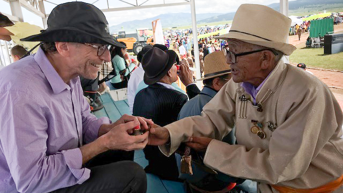 Two men wearing hats sit across from each other smiling and grasping hands.