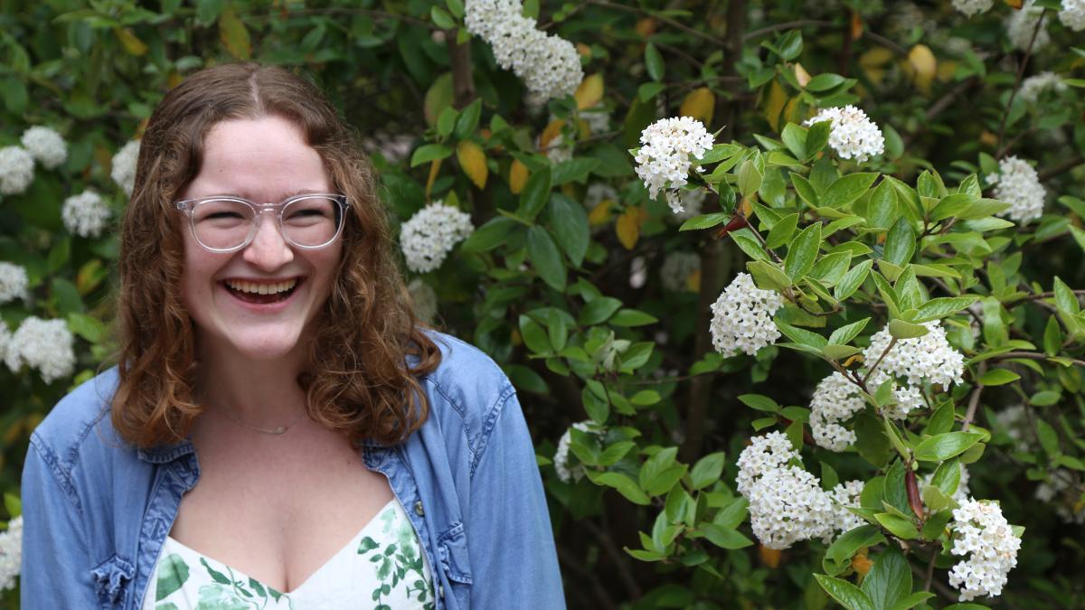 a young woman with glasses wearing a white dress and a blue shirt stands near a flowering bush.