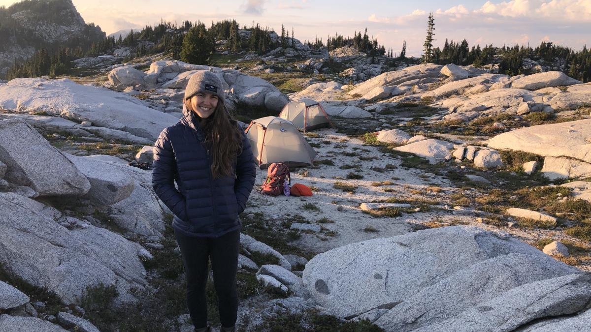 Sam Lovell stands on a rocky outcrop with mountain peaks and tents in the background.