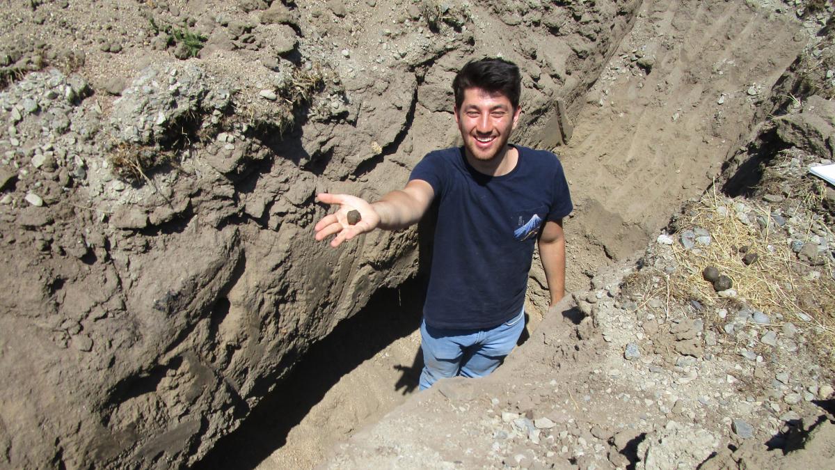 Gombart standing in a hole surrounded by rocks and soil, holding out a ball of soil in his extended right hand.