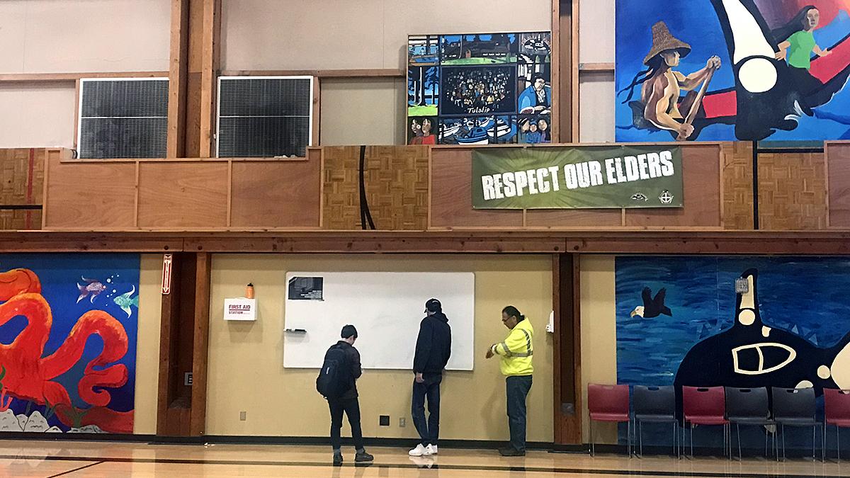 Three people stand in a gym near a whiteboard on a wall decorated with Native American art.