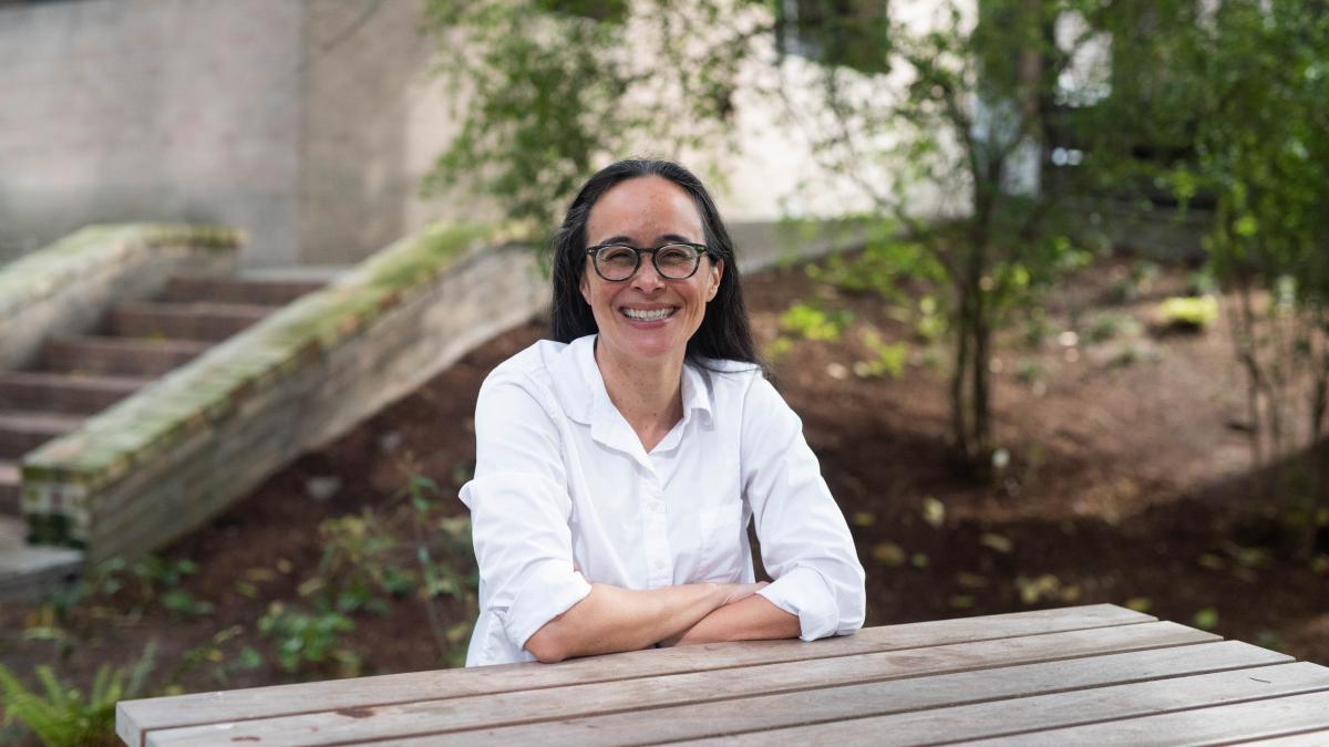 A woman in a white shirt sits outdoors at a picnic table, smiling at the camera.