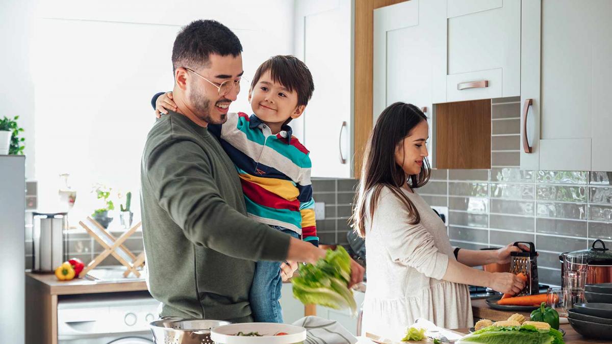 A mid adult Asian couple standing at a kitchen counter in their home, they are preparing a salad for lunch together. The man is preparing lettuce leaves while holding his young son in his arms. His pregnant wife is grating carrots next to him.