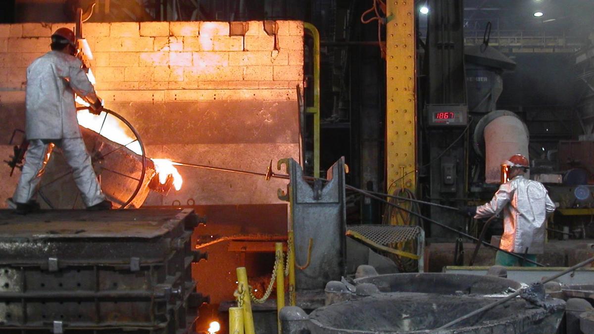 Two workers wear silver protective gear and work in a foundry, one up on a platform over liquid metal which is an orange color. The other is far to the right with a long pole. Both are wearing PPE including helmets and visors.