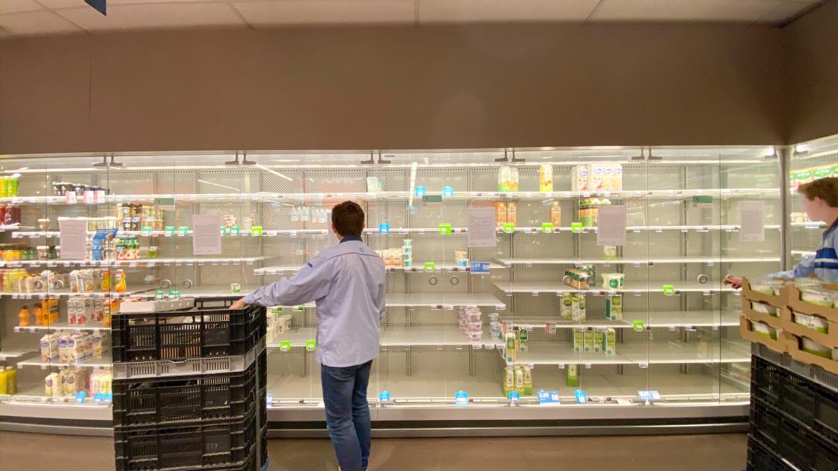 A grocery store employee stands with his back to the camera near a cart of grocery supplies against a backdrop of empty grocery store shelves. 