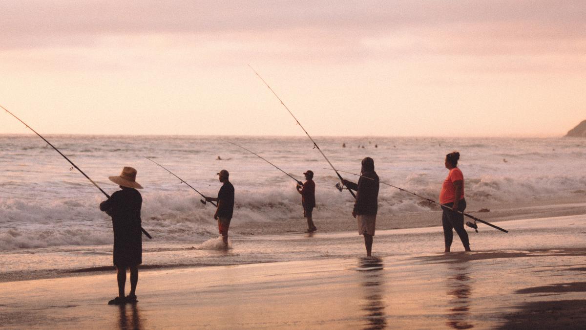 Five people fishing at the ocean, standing in the surf with fishing rods.