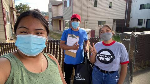 Three people with face masks on stand in front of a fence with houses in background. Person in middle holds a clipboard and pen, person on right gives a peace sign and is wearing a t-shirt that reads "SERVE LOCAL."