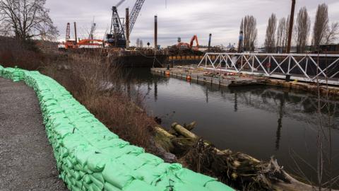 Hundreds of sandbags line a path at the edge of the Duwamish River, with a pier and construction equipment in the background.