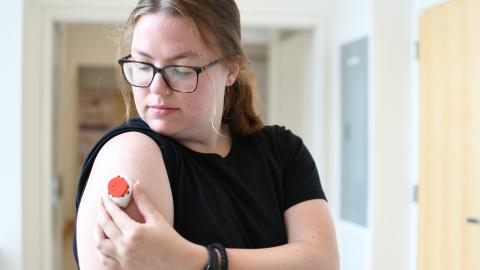 A woman holds a small blood collection device against her upper arm.